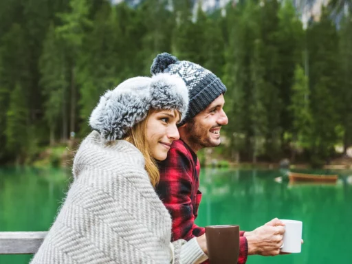 Ein warm gekleidetes Paar sitzt an einem türkisfarbenen See in der Nähe eines Steinmandls, umgeben von Bäumen und Bergen, und hält lächelnd Enzym Komplex Becher in der Hand.