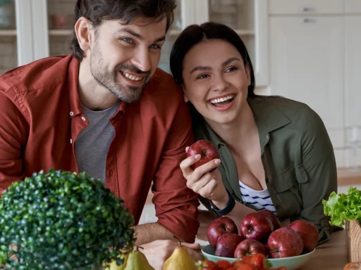 Ein lächelnder Mann und eine lächelnde Frau in einer Küche beugen sich über einen Tisch mit Schalen mit Äpfeln, Erdbeeren, Birnen und einem Granatapfel; eine Flasche OPC Traubenkern Extrakt steht neben einer kleinen Topfpflanze im Vordergrund.