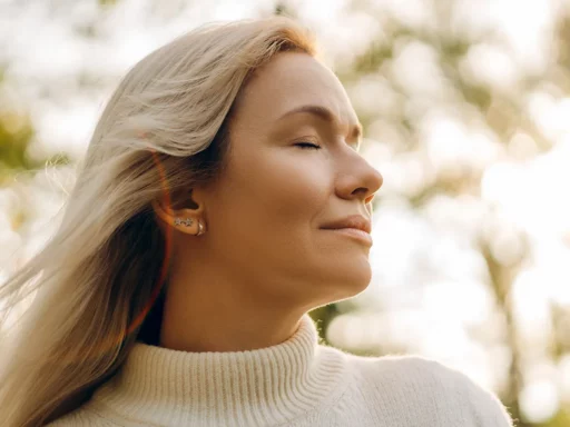 Eine Frau mit langen blonden Haaren und einem cremefarbenen Rollkragenpullover steht im Freien, hat die Augen geschlossen und lächelt friedlich - vielleicht nach der Einnahme von BIO Auricularia Extrakt -, während das Sonnenlicht und die Bäume im Hintergrund sanft verschwimmen.