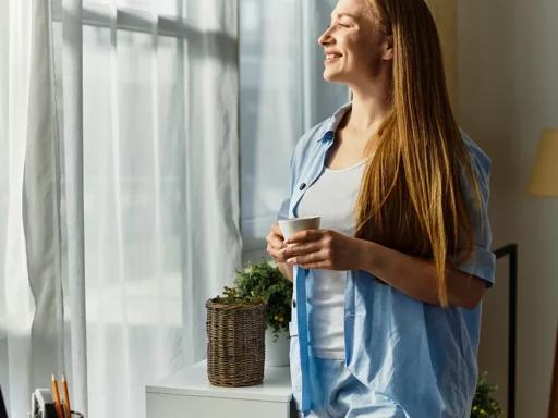 Eine Frau mit langen Haaren, hellblauem Hemd und weißem Top steht lächelnd an einem sonnenbeschienenen Fenster und hält BIO Hericium Extrakt in der Hand. Kopfhörer, Bleistifte und eine Topfpflanze stehen auf dem Schreibtisch neben ihr, während das Sonnenlicht durch die Vorhänge fällt.